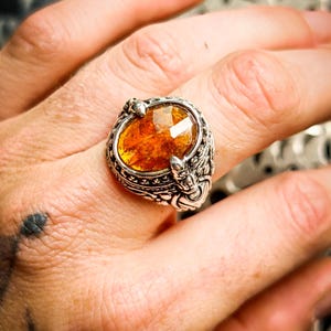 Peut inclure: Une bague en argent ornée d'une pierre facettée de couleur ambre de forme ovale, montée sur une bande métallique détaillée avec des gravures texturées et une finition polie entourant la pierre centrale.