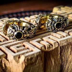 May include: Two ornate gold rings with black center stones rest on a carved wooden surface. The rings feature detailed floral and scrollwork designs. The background is dark, highlighting the rings' intricate details and the wood's texture.