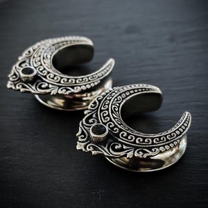 May include: A pair of silver-toned ear plugs featuring a crescent moon shape with intricate embossed scrollwork patterns and a small, round, dark-colored stone set at the center of the outer edge of each piece, showcasing a metallic finish.