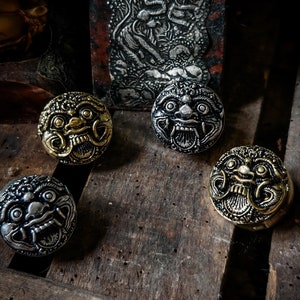May include: Four decorative metal buttons featuring a detailed design of a mythical creature's face. The buttons are in different shades of gold and silver.
