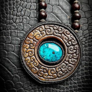 Peut inclure: Un collier avec un pendentif rond en bois sculpté orné d'une pierre turquoise. Le collier est composé de perles en bois brun foncé et est posé sur un fond texturé noir.