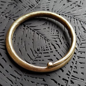 Könnte beinhalten: Ein goldfarbenes schlangenförmiges Armreif-Armband mit strukturierter Oberfläche.
