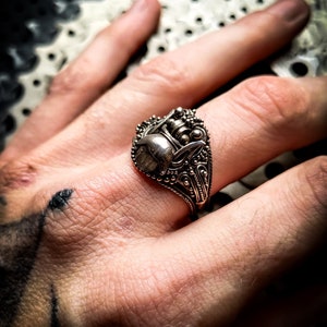 May include: A silver ring with an intricate design featuring a stylised animal head. The ring is worn on a person's finger.
