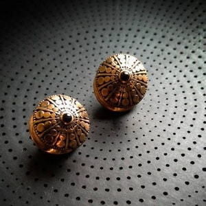 Peut inclure: Deux boucles d'oreilles dorées avec une pierre noire au centre. Les boucles d'oreilles ont un design circulaire avec un motif en relief.