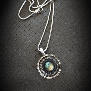 May include: Silver pendant necklace with a round, ornate pendant. The pendant features a central, iridescent stone surrounded by a black border and a detailed silver frame. The chain is a delicate silver snake chain.