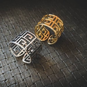 Peut inclure: Deux bagues ouvertes avec un motif géométrique. L'une est de couleur argentée, l'autre dorée. Les bagues sont posées sur un fond sombre et texturé, créant un contraste qui met en valeur leurs motifs complexes. Les bagues sont probablement réglables.