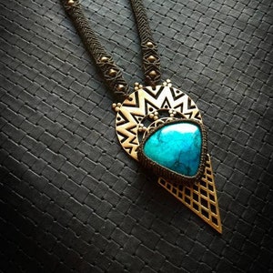 Peut inclure: Un collier avec un pendentif en pierre turquoise et un motif géométrique en laiton. Le collier est fait de cordon noir et a des accents dorés.