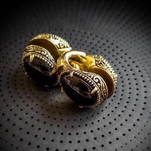 Peut inclure: Deux boucles d'oreilles dorées avec une pierre sombre de forme ovale au centre. Les boucles d'oreilles présentent un motif gravé orné sur les bords. Le fond est une surface sombre et texturée.