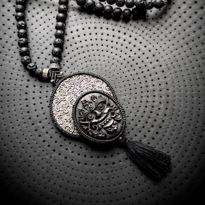 Peut inclure: Un collier de perles noires avec un pendentif argenté et noir représentant un visage sculpté. Le collier est sur un fond noir à motif de points.