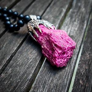 Peut inclure: Un collier composé d'un pendentif en cristal brut magenta vif, enchâssé dans une monture en métal argenté en forme de griffe. Le pendentif est relié à un cordon de perles sphériques noires mates et lisses qui s'étendent vers le haut.