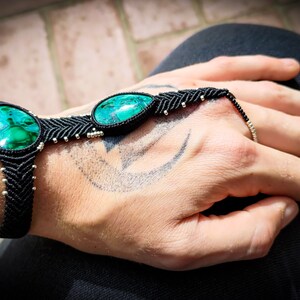 May include: A black macrame bracelet with two green gemstone beads. The bracelet is tied around a person's wrist and has a finger loop.
