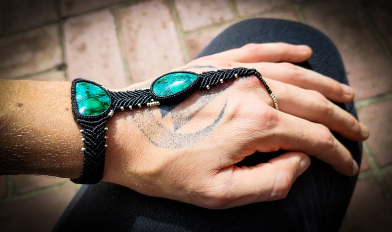 May include: A black macrame bracelet with two green gemstone beads. The bracelet is tied around a person's wrist and has a finger loop.