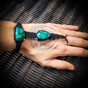 May include: A black macrame bracelet with two green gemstone beads. The bracelet is tied around a person's wrist and has a finger loop.