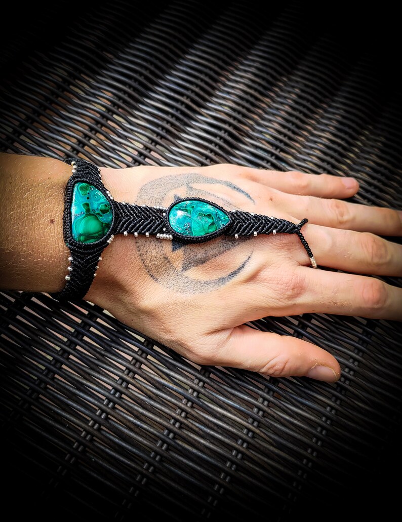 May include: A black macrame bracelet with two green gemstone beads. The bracelet is tied around a person's wrist and has a finger loop.