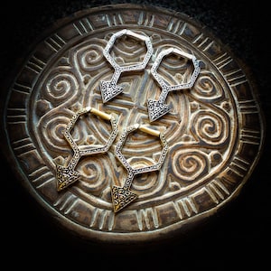 Puede incluir: Cuatro pendientes de aro geométricos en oro y plata con diseños intrincados. Los pendientes están dispuestos sobre una superficie de madera con un patrón tallado.