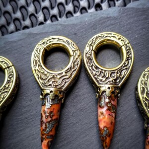May include: Three gold-toned metal ear gauges with intricate designs and a pointed, red and white stone attached to the bottom.