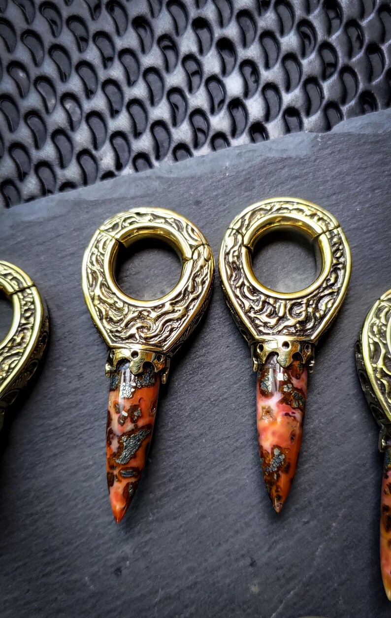 May include: Three gold-toned metal ear gauges with intricate designs and a pointed, red and white stone attached to the bottom.