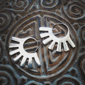 May include: A pair of silver hoop earrings with a sunburst design. The earrings are on a patterned surface.
