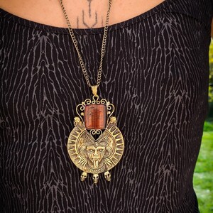 May include: A gold necklace with a large pendant featuring a detailed, ornate design. The pendant has a red gemstone in the center and three skull-shaped charms hanging from the bottom.
