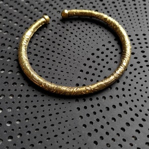 Könnte beinhalten: Ein goldfarbenes Armreif-Armband mit einem filigranen Blumenmuster. Das Armband liegt auf einer schwarzen Oberfläche mit einem Muster aus kleinen Löchern.