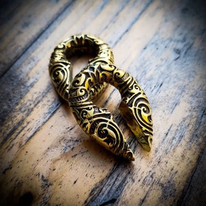 May include: Two gold-colored metal ear gauges with intricate floral designs. The gauges are shaped like a ring with a pointed end.