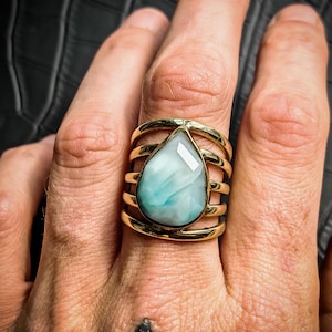 Peut inclure: Une bague dorée ornée d'une pierre précieuse bleu clair en forme de goutte d'eau. La bague présente un design à plusieurs bandes. La pierre est sertie dans une lunette dorée. La bague est sur un doigt, avec un fond noir.