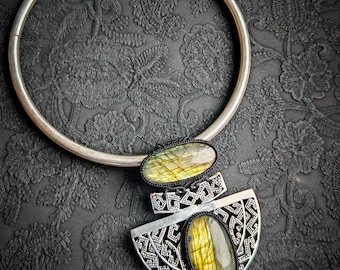 Puede incluir: Un collar plateado con un colgante grande con dos piedras preciosas ovaladas e iridiscentes. El colgante tiene un diseño geométrico. El collar se exhibe sobre una superficie negra texturizada.