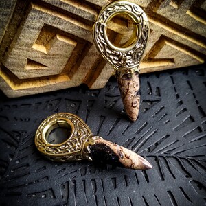 May include: A pair of gold-toned metal ear gauges with a decorative design and a brown and white stone tip.