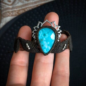 Puede incluir: Un brazalete de piedra turquesa con un engaste de plata y un cordón negro. El brazalete está atado alrededor de una mano.