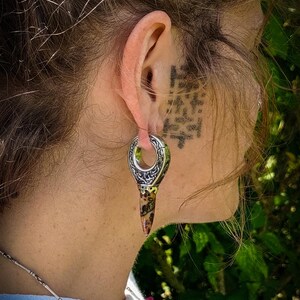 May include: A silver and brown ear gauge with an intricate design. The gauge is worn in the ear of a person with brown hair.
