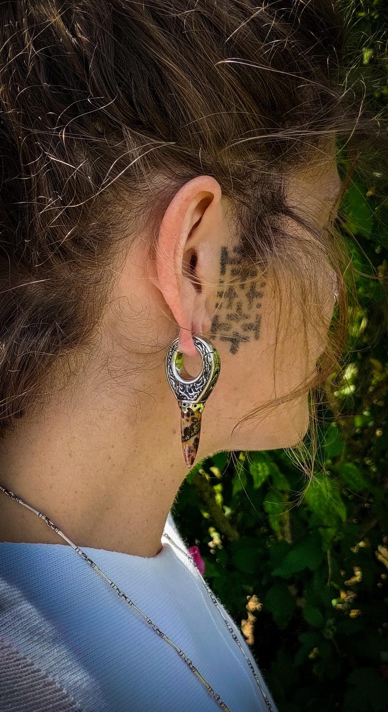May include: A silver and brown ear gauge with an intricate design. The gauge is worn in the ear of a person with brown hair.