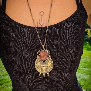 May include: A gold necklace with a large, ornate pendant featuring a red stone and a stylized face. The pendant is surrounded by a circular design with skulls at the bottom.