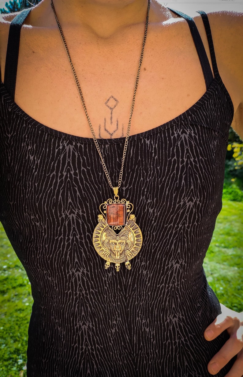 May include: A gold necklace with a large, ornate pendant featuring a red stone and a stylized face. The pendant is surrounded by a circular design with skulls at the bottom.