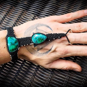 May include: A black macrame bracelet with three green gemstone beads. The bracelet is worn on a person's wrist and hand, with the finger loop extended.
