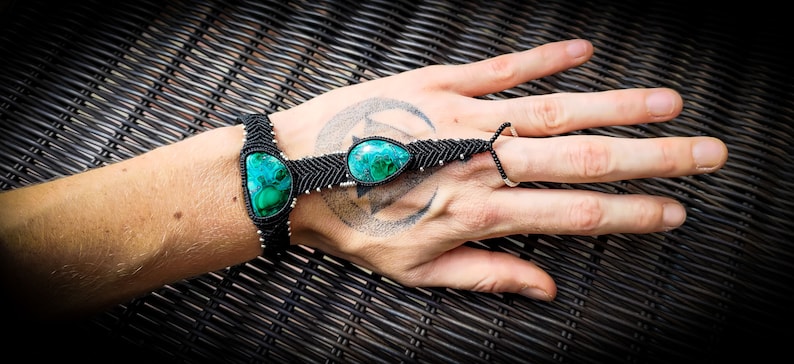May include: A black macrame bracelet with three green gemstone beads. The bracelet is worn on a person's wrist and hand, with the finger loop extended.