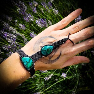 May include: A black macrame bracelet with two green gemstone accents. The bracelet is tied around a wrist and has a finger loop.