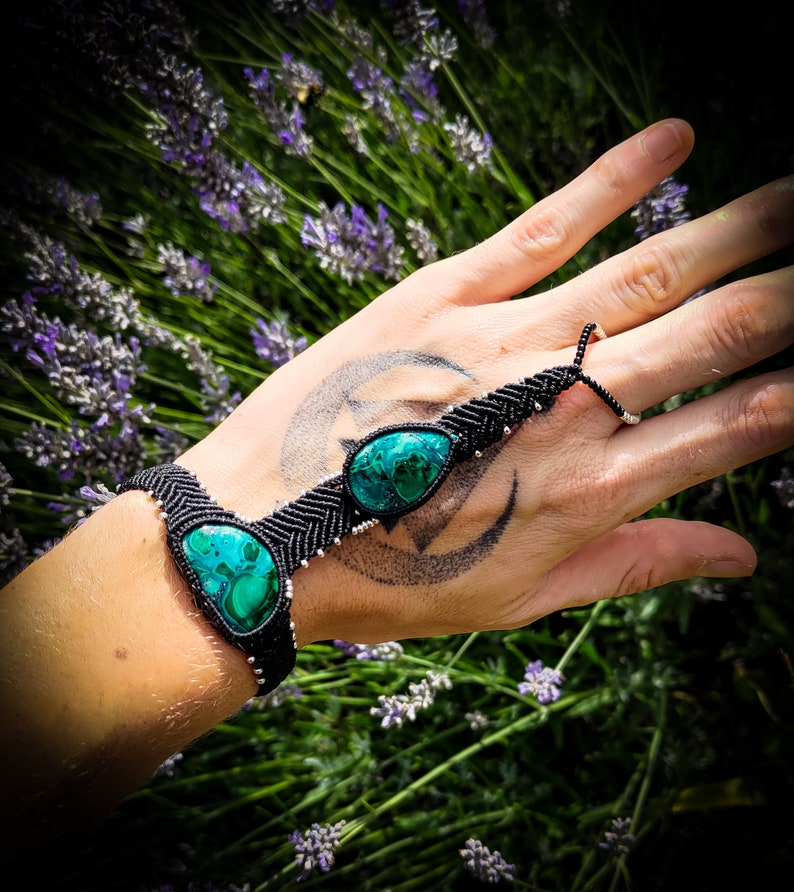 May include: A black macrame bracelet with two green gemstone accents. The bracelet is tied around a wrist and has a finger loop.