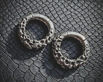May include: Two silver hoop earrings with a textured, organic design. The earrings are on a black, textured surface.