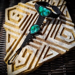 May include: Black macrame bracelet with two green malachite stones. The bracelet is on a wooden surface with a geometric pattern.