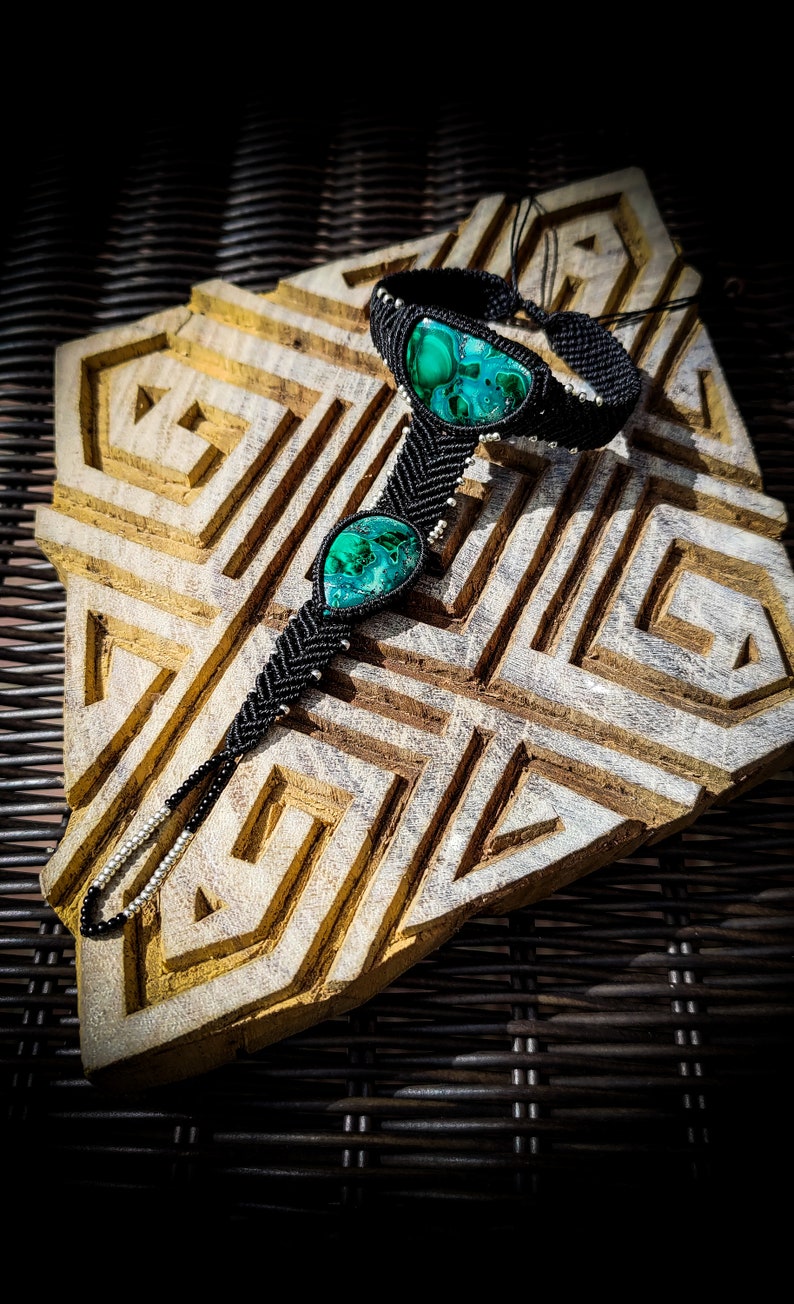 May include: Black macrame bracelet with two green malachite stones. The bracelet is on a wooden surface with a geometric pattern.