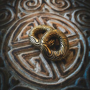 Peut inclure: Deux anneaux en métal couleur or avec un motif texturé en spirale. Les anneaux sont sur une surface à motifs brun et bleu.