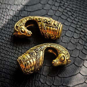 Puede incluir: Dos objetos ornamentales de metal dorado con intrincadas tallas y una forma curva. Cada pieza presenta una cabeza de animal estilizada. Se exhiben sobre una superficie oscura y texturizada, creando un contraste llamativo.