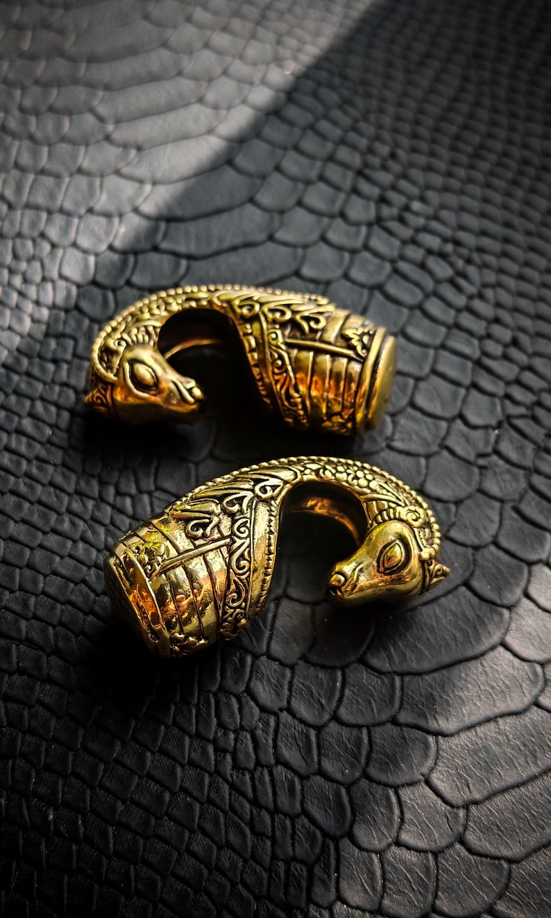 Puede incluir: Dos objetos ornamentales de metal dorado con intrincadas tallas y una forma curva. Cada pieza presenta una cabeza de animal estilizada. Se exhiben sobre una superficie oscura y texturizada, creando un contraste llamativo.