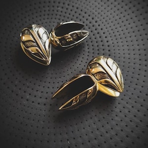May include: Two ornate metal ear gauges, one gold and one silver, with intricate designs. The gauges are on a black textured surface.