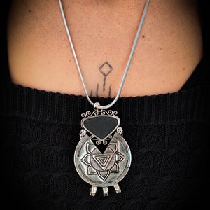 May include: A silver necklace with a black stone pendant. The pendant is a circle with a triangle in the center and a smaller triangle inside the larger triangle. The pendant is surrounded by a decorative design.