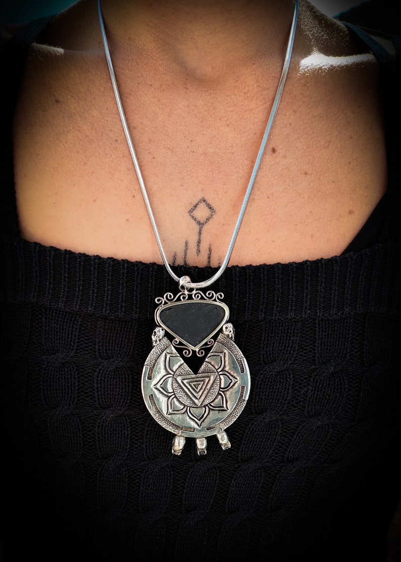 May include: A silver necklace with a black stone pendant. The pendant is a circle with a triangle in the center and a smaller triangle inside the larger triangle. The pendant is surrounded by a decorative design.
