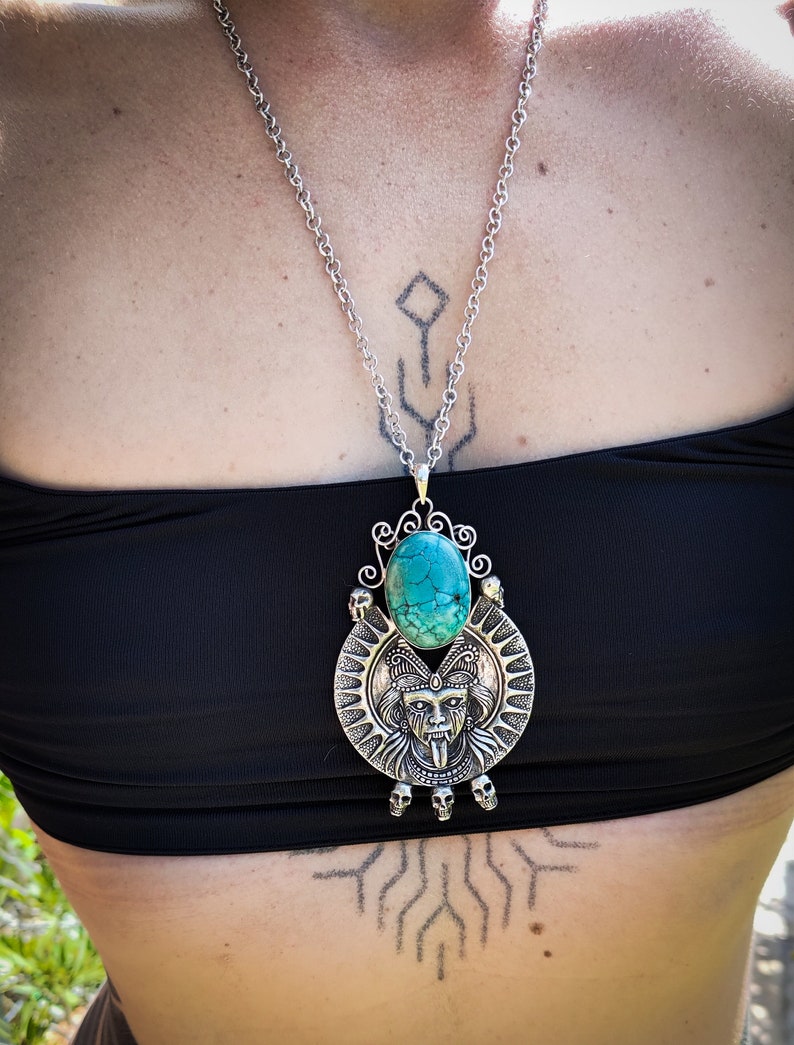 May include: A silver necklace with a turquoise stone pendant featuring a detailed skull and face design. The pendant is surrounded by smaller skulls.