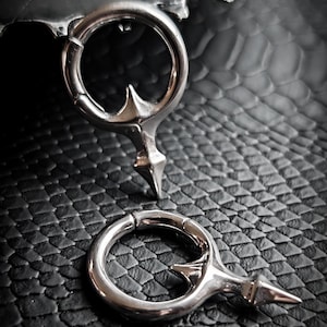 Puede incluir: Dos pendientes de aro plateados con un diseño puntiagudo. Los pendientes están colocados sobre un fondo oscuro y texturizado. Los pendientes tienen un estilo único y atrevido.