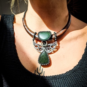 Peut inclure: Un collier ornemental avec une base en forme de croissant en bois foncé. Le collier présente un pendentif argenté avec des pierres vertes. Le pendentif a un motif d'ailes d'ange avec une pierre verte triangulaire en bas.