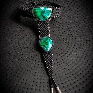 May include: A black macrame ankle bracelet with two green malachite stones. The bracelet is tied with a knot at the ankle and has silver beads throughout.